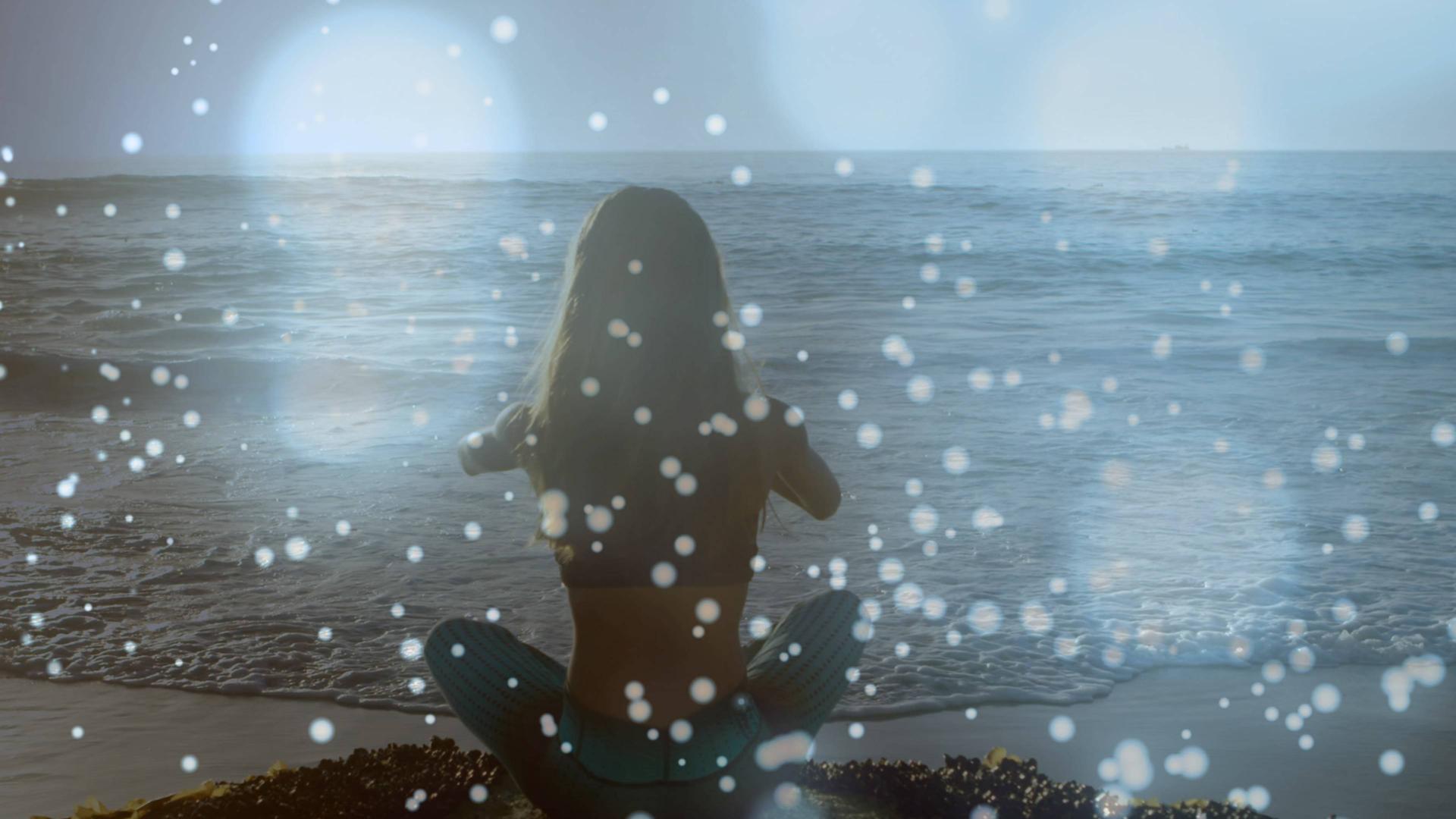Eine Person in Yogapose sitzt dem Betrachter mit dem Rücke zugewandt am Meer. Über dem Bild liegt eine Fläche aus mystischem Schillern, was dem Bild eine spirituelle Stimmung verleiht. Eine Person in Yogapose sitzt dem Betrachter mit dem Rücke zugewandt am Meer. Über dem Bild liegt eine Fläche aus mystischem Schillern, was dem Bild eine spirituelle Stimmung verleiht.