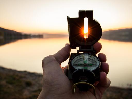 Ein Kompass in der Hand zeigt Richtung Westen, auf die untergehende Sonne.